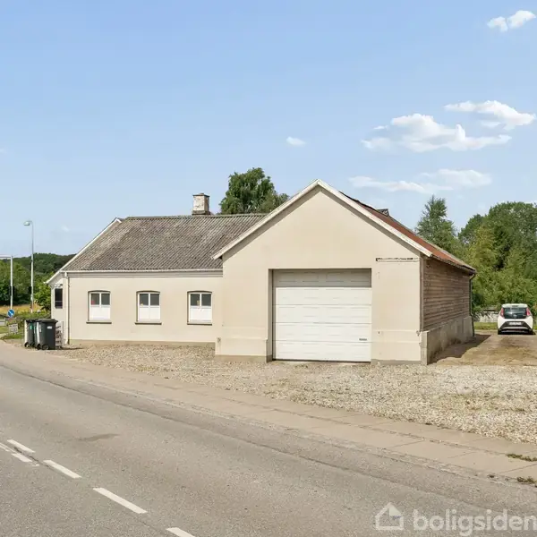 En lys beige bygning med garageport står ved en landevej. Bygningen er omgivet af grus og asfaltveje med skovområde i baggrunden. En hvid bil ses ved siden af.