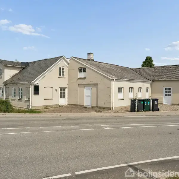 Et stort, beige hus står stille ved en vej. Huset har flere sektioner med forskellige tagudhæng. Tre affaldscontainere står foran huset, omgivet af en grusbelagt gård.