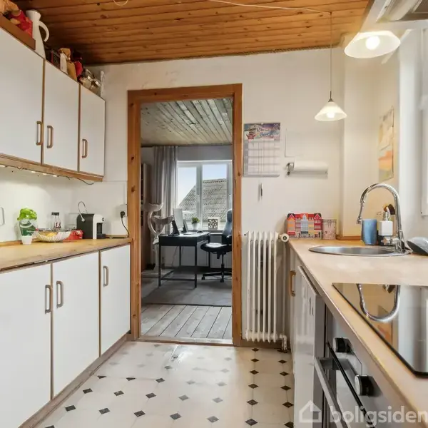 Køkkenet har hvide skabe og træbordplade, med en kaffemaskine og elkedel på bordet. En dør fører til et tilstødende rum. Rummet har hvide vægge og sort/hvidt ternet gulv.