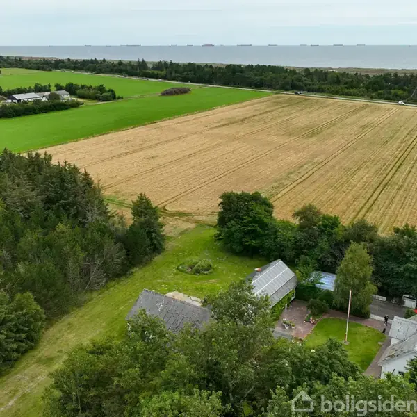 Et hus, markeret med en rød pil, ligger blandt træer og marker nær kysten. Omkringliggende landbrugsmarker strækker sig til horisonten, med havudsigt i baggrunden.