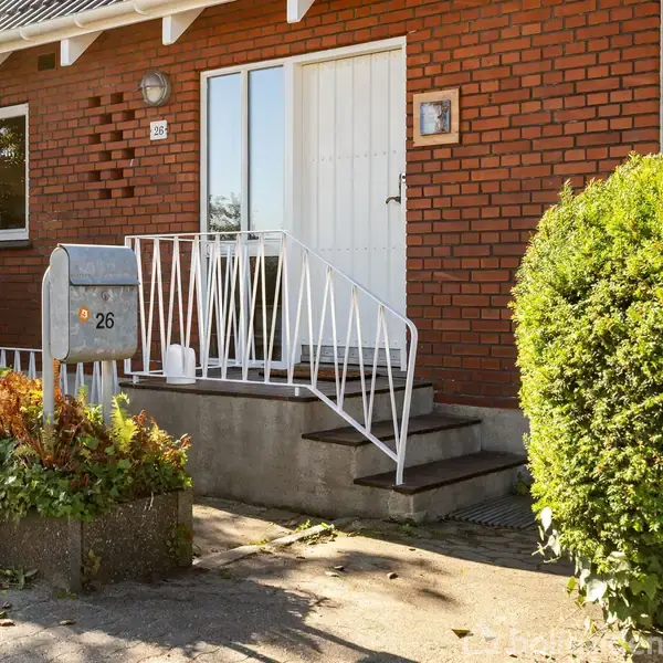 Rød murstenshus med hvid hoveddør og trappe. En postkasse med tallet 26 står ved siden af indgangen. Omgivet af grønne buske og en stenbelagt gangsti.