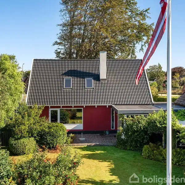 Rødt hus med sort tag, stående i en frodig have med flagstang og tæt omkranset af grønne buske og træer. Klar blå himmel som baggrund.