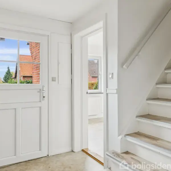 En indgang med en hvid dør til venstre, lys, åben udsigt udenfor. En trappe til højre fører opad. Lyse vægge og minimalistisk design præger rummet.