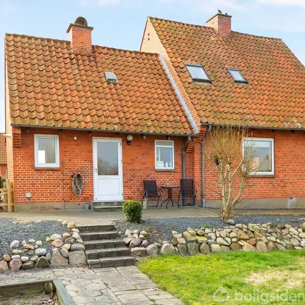 Et rødt murstenshus med en brun tegltag sidder i en have. Stenen er stablet langs bunden, en trappe fører til indgang, mens stole og et bord står udenfor.