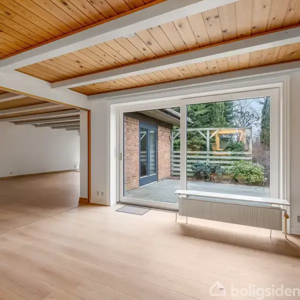 En lys stue med trægulv og træloft. En stor glasdør leder ud til en terrasse omgivet af buske og træer. En radiator står under vinduet.