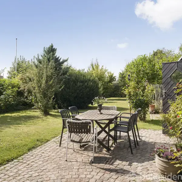 Et udendørs spisebord med seks stole står på en brostensbelagt terrasse. Bordet har en blomstervase. Omgivet af velplejet græsplæne og frodig bevoksning under en klar blå himmel.