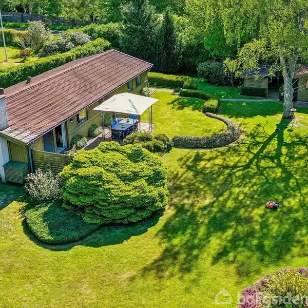 Hus med skrå tag og skorsten, omkranset af velplejet have med buske og træer. Parasol og havemøbler står på terrassen. Grønne omgivelser skaber fredfyldt atmosfære.