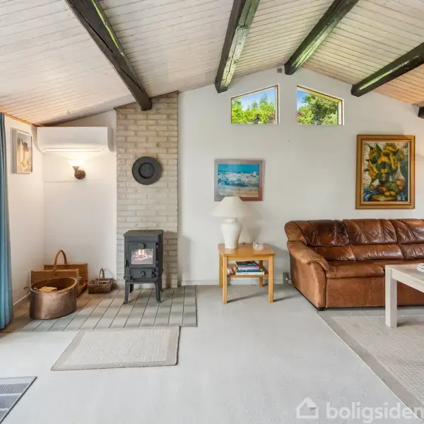 En stue med brændeovn og lys murstensvæg. Brune lædersofaer og et sofabord står i rummet. Store vinduer med gardiner og træpaneler skaber en lys og hjemlig atmosfære.