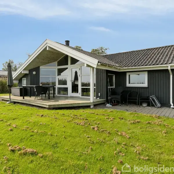 Et mørkt træsommerhus med store vinduer og terrasse, står på en frodig græsplæne. Terrassen har et bord med stole og en parasol. Omkring er grønne træer og hække.