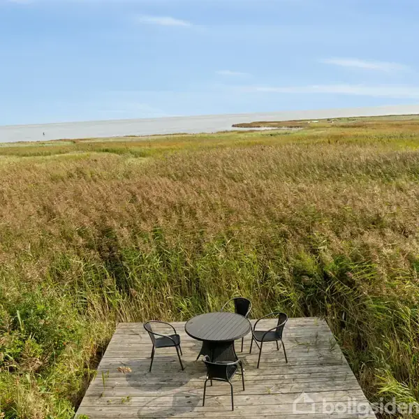 En bord-og-stolesæt står på en træterrasse omgivet af høje siv og græs med udsigt over en fjern sø eller hav i horisonten under en blå himmel.