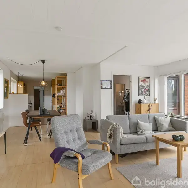 En stue med gråt sofaarrangement og lænestol står ved et sofabord. Spisebord med stole ses i baggrunden. Store vinduer giver lys og udsigt til røde murstensbygninger.