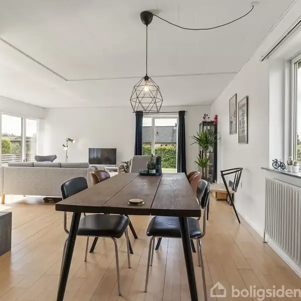 Et træbord med sorte stole står i en spisestue med lyse trægulve. En moderne lampe hænger over bordet. I baggrunden ses en stue med sofa og fjernsyn.