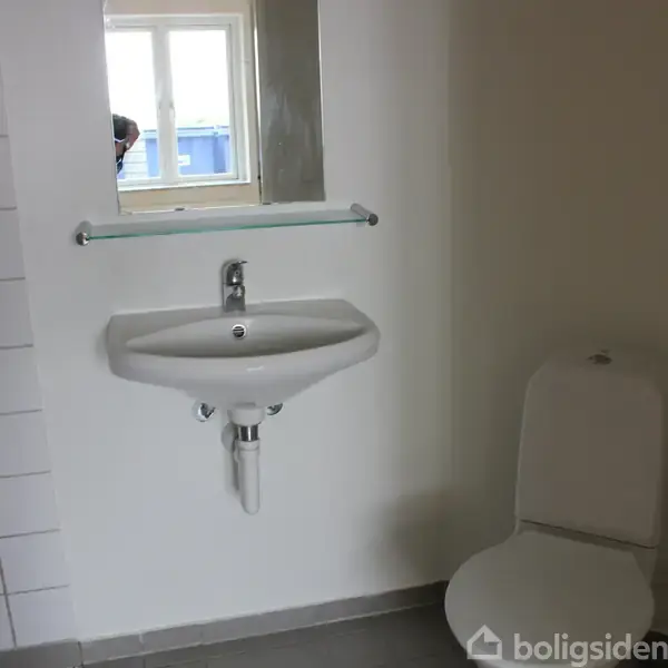 Håndvask hænger på væggen over toilettet i et badeværelse. Håndvasken er under et spejl og ved siden af et vindue. Gulvets farve er grå.