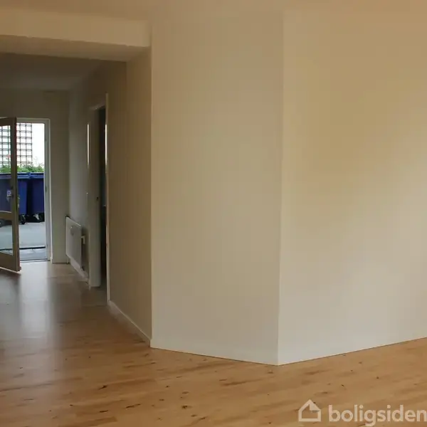 Et lyst rum med trægulv fører til en åben dør mod en balkon. Til venstre ses et skab og en gang. Væggene er lyse.
