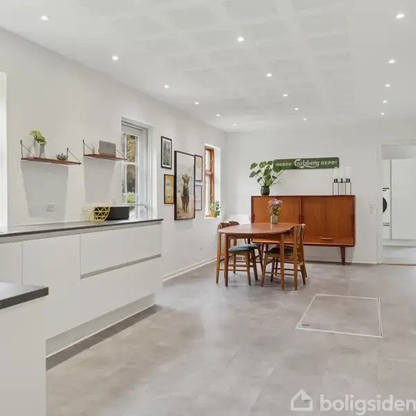 Et moderne køkken med hvide skabe og sorte bordplader ved siden af en træspisebord med stole. Store vinduer giver lysindfald. Dekorationer på hylderne og lyse vægge fuldender rummet.