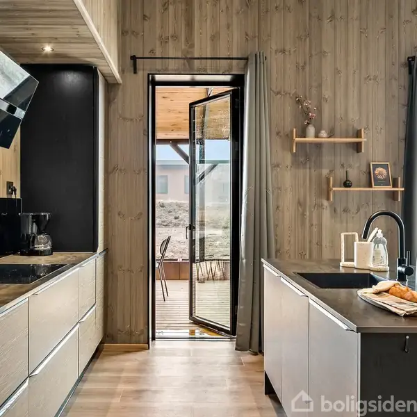 Et moderne køkken med lyse træskabe, en køkkenø og en sort vask. En åben dør leder ud til en terrasse. Væggene er beklædt med træpaneler.