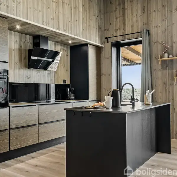 Køkken med moderne apparater; ovn, emhætte og køleskab indbygget i træborde. Køkkenø med vask og vandhane. Store vinduer og trævægge skaber en lys, naturlig atmosfære.