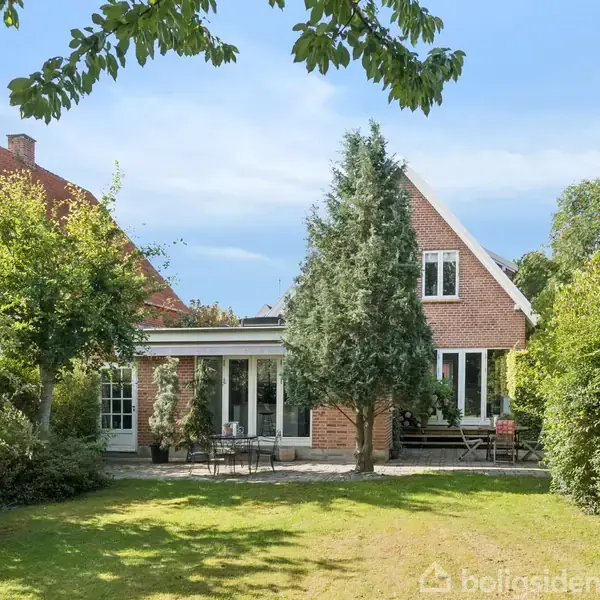Huset står stille omgivet af en grøn have med en terrasse foran. Røde murstensvægge og skrå tagflader med vinduer præger huset. Skyfri himmel tilføjer lys.
