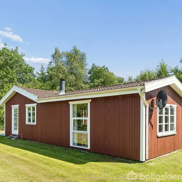 Rødt sommerhus med hvide vinduer står på en velplejet græsplæne omgivet af træer under en blå himmel med enkelte skyer. En satellitdisk er monteret på husets side.