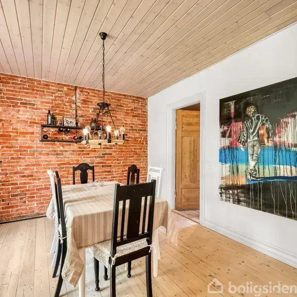 Spisebord med seks stole står i en spisestue med en murstensvæg. En lysekrone hænger over bordet, og et maleri hænger på den hvide væg ved siden af en trædør.