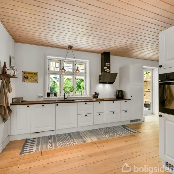 Køkken med hvide skabe og integrerede apparater står ubenyttet. Sollys strømmer ind gennem et vindue over vasken. Der er en åben dør til en terrasse udenfor.