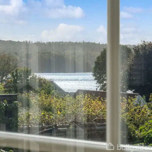 Udsigt gennem et vindue, hvor sollys glitrer på en sø omgivet af frodige træer og buske. I baggrunden ses en skov under en blå himmel med lette skyer.
