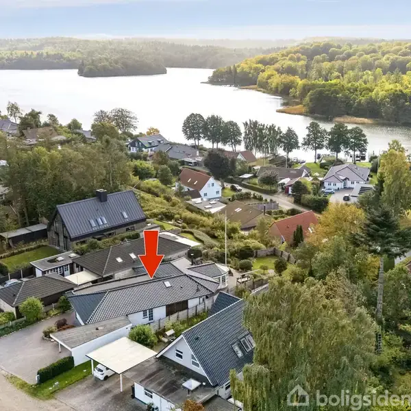 Et parcelhus set oppefra, markeret med en rød pil, ligger i en lille, skovklædt by nær en sø.