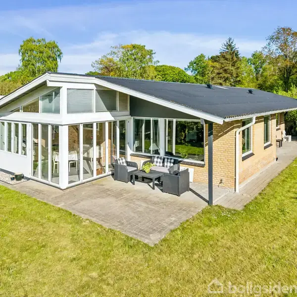 Et enfamiliehus med store vinduer og terrasse møbleret med sofa og bord, beliggende i en grøn have omkranset af træer under en klar blå himmel.