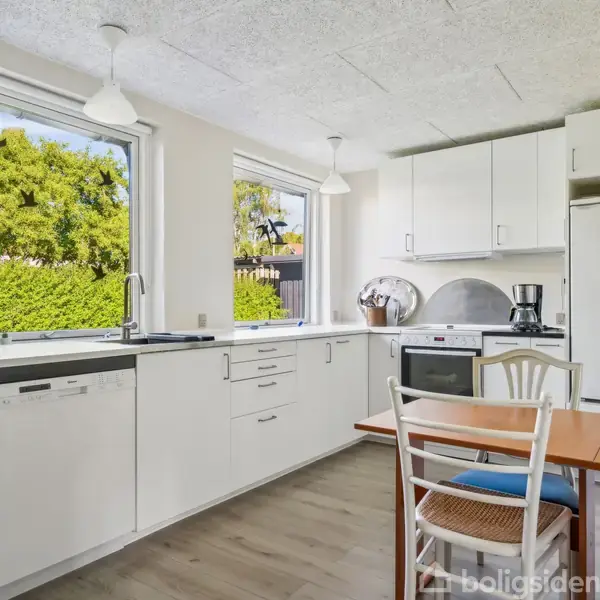 Køkken med hvide skabe og apparater, hvor et bord og stole er placeret ved siden af et køleskab. Store vinduer giver lys, omgivet af grønne buske udenfor.