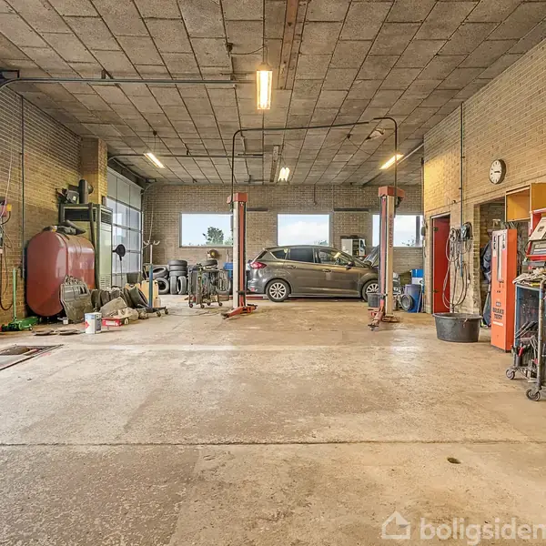 En bil holder på en autolift i en garage med værktøj og udstyr spredt rundt. Der er store vinduer og en åben garageport, der slipper lys ind. En væg har en urskive.