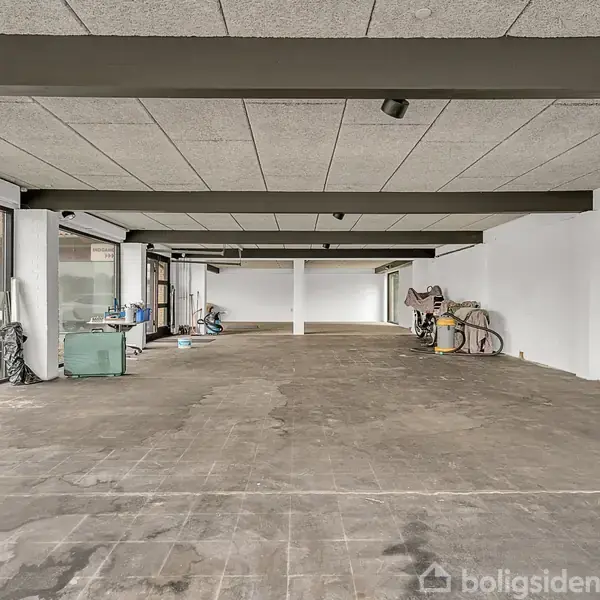 Stor, tom garage med store vinduer, betonloft og gulv; rengøringsartikler og bilværktøj langs væggen; parkerede biler udenfor gennem vinduet.