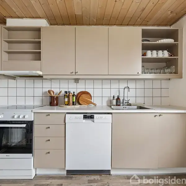 Køkken med beige skabe over en hvid flisevæg, en ovn, opvaskemaskine og vask. Bordpladen er hvid, og der er pynteplanter samt køkkenredskaber på bordet.