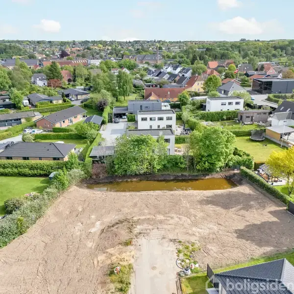 Bebyggede parcelhusområder omgiver en åben jordgrund med en lille dam og spredte træer, omkranset af buske og hegn, i et frodigt, forstadsagtigt kvarter.