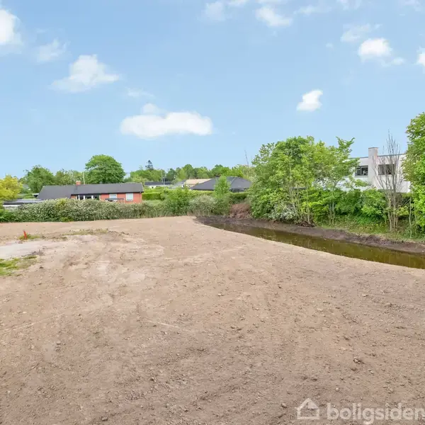 Et åbent, jordet område ligger stille, omgivet af grøn vegetation og huse i baggrunden, under en blå himmel med lette skyer.