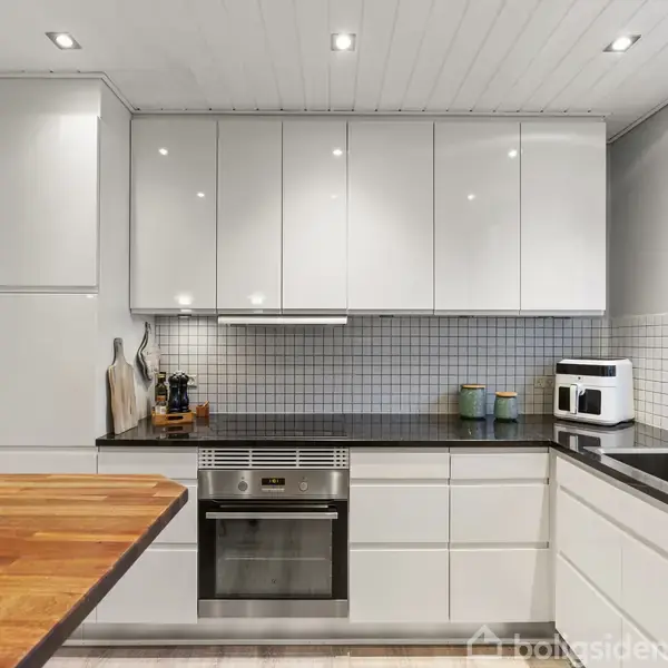 Et moderne køkken med hvide skabe, en indbygget ovn og sort bordplade. Der er en stor træbordplade til venstre. Vinduet til højre giver udsigt til en have.