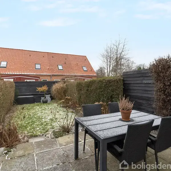 Et bord med fire stole står på en terrasse. En plantepotte pynter bordet. Omkring terrassen er der hække, og i baggrunden ses et hus med rødt tegltag.