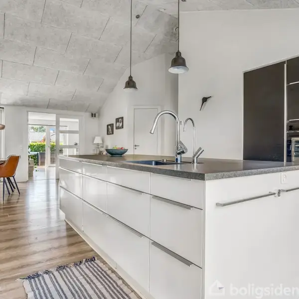 Køkkenbord med håndvask og mørk bordplade i et moderne køkken. Bagved ses spisebord med orange stole, store vinduer og lysindfald, samt trægulve og grå loftslamper.