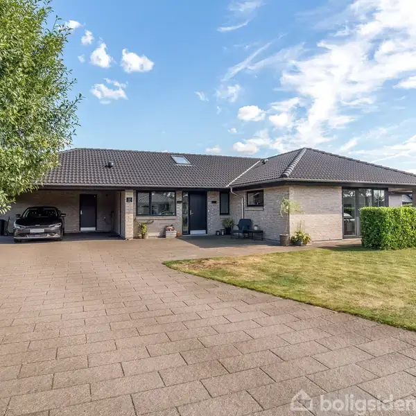 Et moderne hus med en mørk tag, omgivet af en velplejet græsplæne og flisebelagt indkørsel. Træer står tæt ved siden af, og en bil er parkeret i carporten.