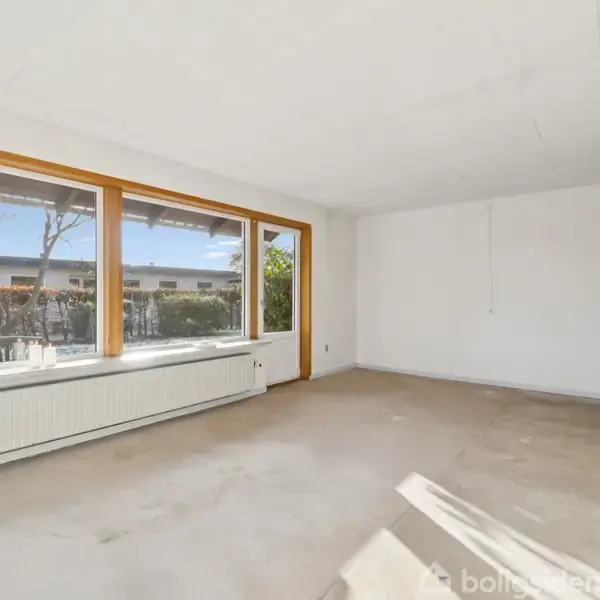 Et tomt lokale med store vinduer, der slipper lys ind. En radiator og en plante står under vinduerne. Væggene er hvide og gulvet er lyst, uden møbler.