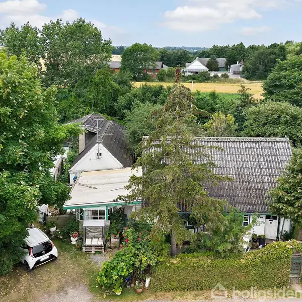 Hus med stråtag omgivet af træer. Foran huset står en lille hvid bil. Landskabet omkring er grønt med marker og spredte bygninger i baggrunden.