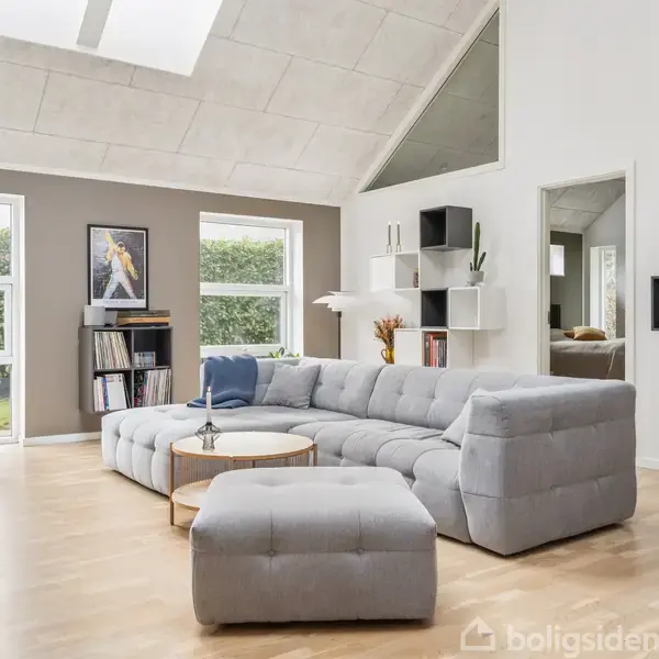 Stor grå sofa i moderne stue, placeret på trægulv med minimalistiske hylder. Store vinduer med udsigt til en hæk, skaber et lyst rum. Maleri og planter tilføjer dekoration.