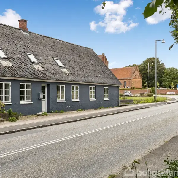 Et langstrakt, blåmalet hus med mange vinduer står langs en vej. Huset har et mørkt skrått tag med ovenlysvinduer. Omkringliggende har en landsbyagtig atmosfære med veje og grønne områder.