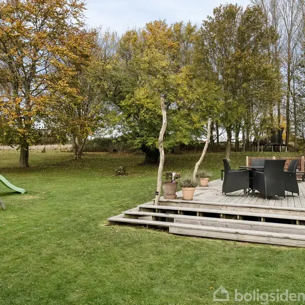 En træterrasse med sorte møbler og blomsterkrukker står i en have. Ved siden af er en gyngestativ med rutsjebane. Omkring er græsplæne og træer.