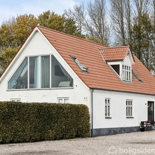 Hvidt hus med rødt tag står stille på grusvej, omgivet af buske og træer i efterårsfarver. Øverst ligger et stort trekantet vindue.