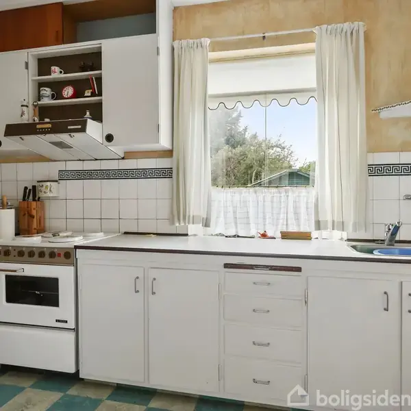 En hvid køkkenvask er placeret under et vindue med hvide gardiner, ved siden af en komfursektion. Køkkenskabe omgiver området med blå-hvidt ternet gulv og vægfliser med mønster.