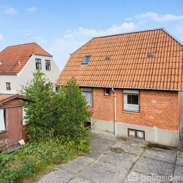 Et rødt murstenshus med rødt tegltag står i et boligområde. Foran huset er der en grusbelagt indkørsel, en træskur og grøn beplantning. Himlen er klar blå.