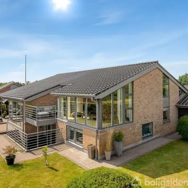 Et moderne to-etagers hus med store vinduer og en veranda. Det står i en grøn have med velplejet græsplæne og buske. Blå himmel og solskin i baggrunden.