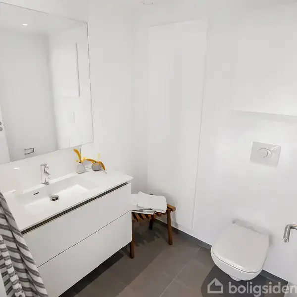 Badeværelse med hvidt skabsarrangement under vask, spejl ovenfor, toilet på højre væg, håndklædeholder ved siden af, og en grå flisebelagt gulv i lys indretning.