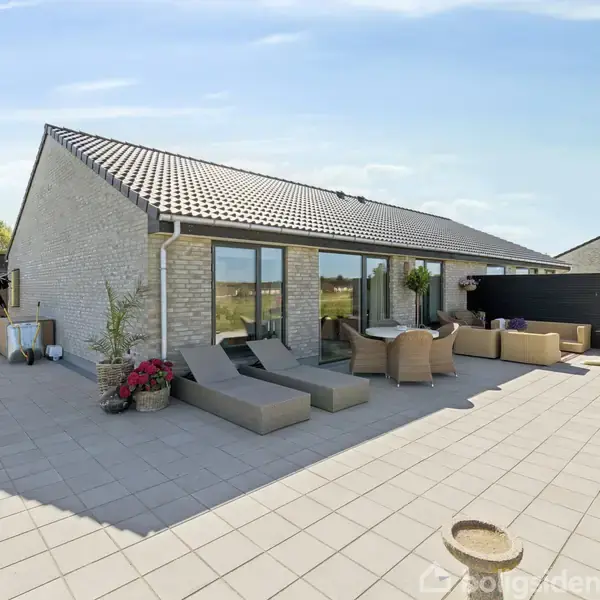 Moderne hus med fladt tag, store vinduer og en stor terrasse. Terrassen har loungemøbler, liggestole og planter. I baggrunden ses en græsplæne og en klar blå himmel.