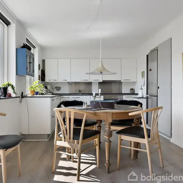 Et rundt bord med fire træstole står i et lyst køkken. En lampe hænger over bordet, og der er planter i vindueskarmen. Møbler og køkkenelementer er moderne.